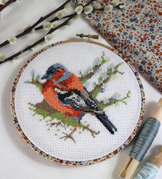Cross-stitched piece of a bird on a branch with embroidery tools and fabric in the background.