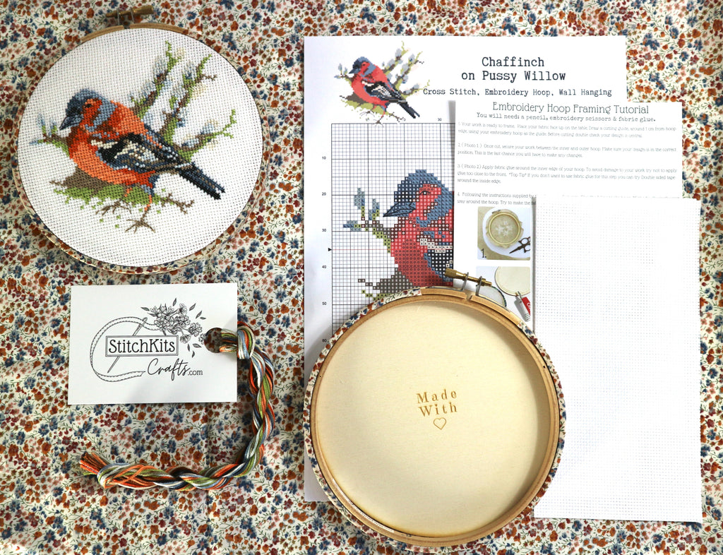 Embroidery kit with cross-stitched bird design, hoop, and pattern sheet on a floral fabric background.