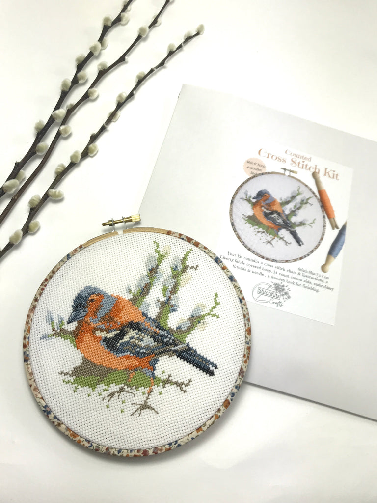 Cross-stitched bird design in an hoop with kit packaging on a white background