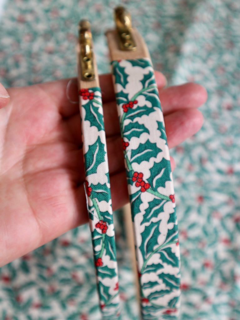 Decorative embroidery hoops with holly pattern held by a hand against a floral background
