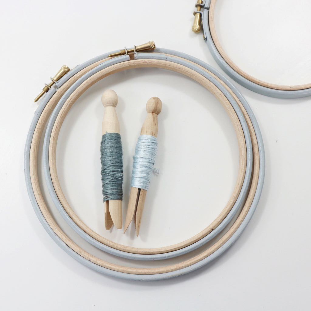 Pale gray embroidery hoops with embroidery threads on peg dolls.