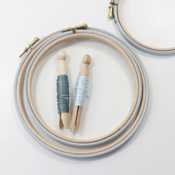 Pale gray embroidery hoops with embroidery threads on peg dolls.